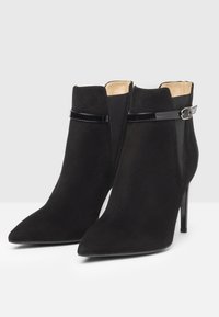 Sorte ruskindsankelstøvler med spids tå, stilettohæl og blank accentrem detaljeret ved anklen.