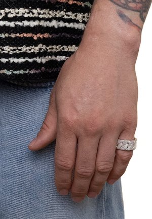 Silver ring with a braided design on the index finger; textured surface; hand resting against denim fabric with light blue color.