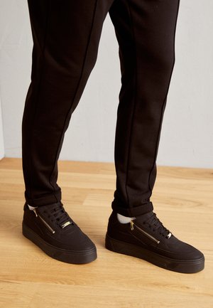 Person trägt schwarze Sneakers mit goldenen Reißverschlüssen und schwarze Hose und steht auf einem hellen Holzboden.