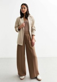 Blazer de lana beige sobre una blusa marrón de canalé, combinado con pantalones marrones de pierna ancha y zapatillas blancas. Predominan las texturas planas y suaves.