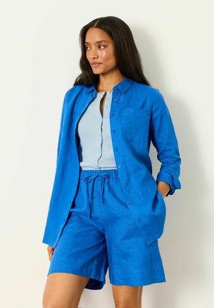 Frau mit langen, dunklen Haaren, die ein blaues Hemd mit Knöpfen und passende Shorts über einem hellblauen Top trägt, steht mit einer Hand in der Hosentasche.