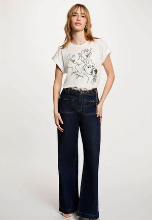 Giovane donna con capelli alla lunghezza delle spalle che indossa una maglietta bianca con grafica e jeans scuri a gamba larga a vita alta con cintura decorativa e catena.