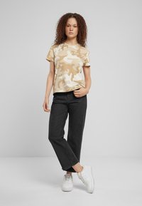 Maglietta a maniche corte in camouflage beige, abbinata a jeans scuri e sneakers bianche, con una vestibilità rilassata e una texture di tessuto morbida.
