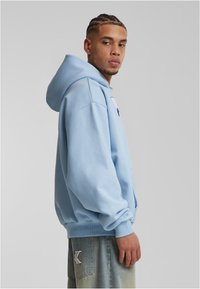 Felpa azzurro chiaro con una vestibilità rilassata, grande tasca frontale e polsini a costine. Abbinata a jeans grigi chiari con logo ricamato.