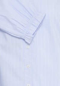 Chemise bleu clair à fines rayures avec manchettes élastiquées froncées et boutons blancs sur la patte de boutonnage avant.