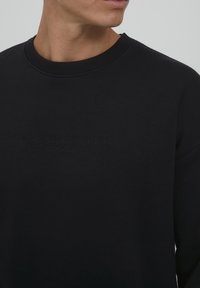 Fekete, pamut crewneck pulóver; texturált bordás nyakkivágással és dombornyomott logóval a frontján.