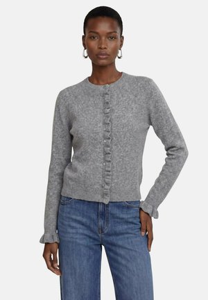 Kvinde iført en grå strikket cardigan med flæset knapstolpe og manchetter, kombineret med blå jeans, stående mod en ensfarvet baggrund.