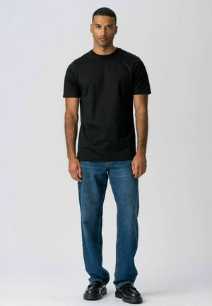 Uomo in piedi contro uno sfondo semplice, indossa una maglietta nera, jeans blu e scarpe nere, guarda dritto con un'espressione neutra.