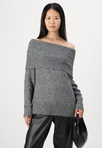 Pull gris à épaules dénudées avec un col en rib et des poignets, associé à un pantalon en cuir noir, maintenu par un sac noir noué.