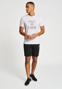 Hvid T-shirt med korte ærmer og et sort "hummel" logo, parret med sorte shorts og sorte sneakers. Outfitet har et minimalistisk design.