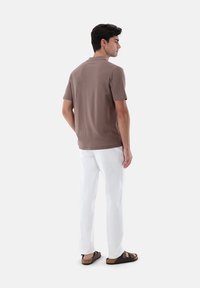 Polo shirt marrone a maniche corte con una texture liscia, abbinato a pantaloni bianchi e sandali marroni, visto da dietro.