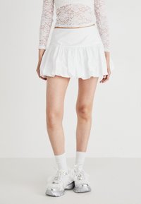 Mini gonna bianca con volant e una texture liscia. Indossata con sneakers chunky argento, top bianco a maniche lunghe in pizzo e calzini bianchi.