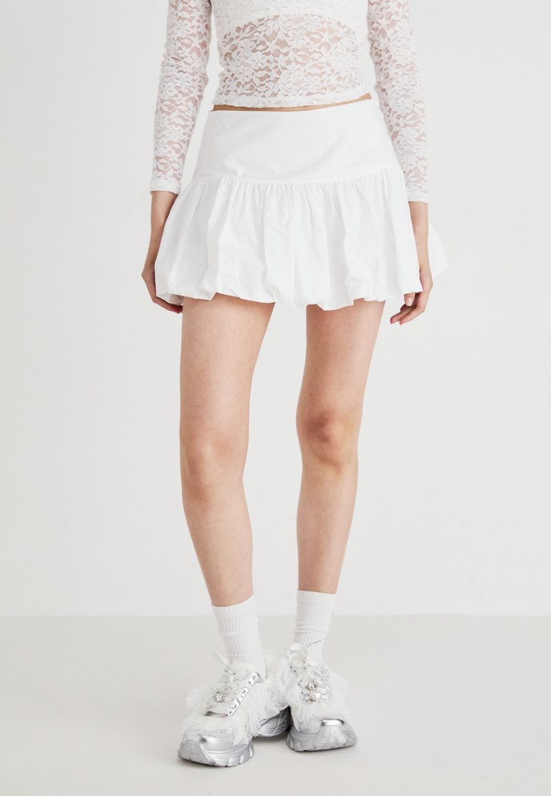 Mini gonna bianca con volant e una texture liscia. Indossata con sneakers chunky argento, top bianco a maniche lunghe in pizzo e calzini bianchi.