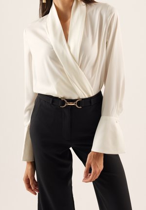 Femme portant un chemisier cache-cœur blanc avec des poignets évasés et un pantalon noir avec une ceinture noire ornée de boucles dorées.