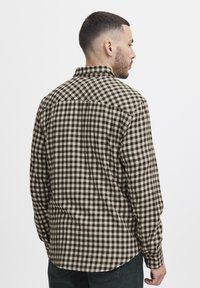 Camisa de franela a cuadros marrón y crema con cuello abotonado, mangas largas y dobladillo curvado. Tela texturizada visible en la parte posterior.