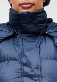 Námorně modrá péřová bunda s vysokým límcem, s hladkou, lesklou texturou, knoflíkovými akcenty a detaily bočních panelů.