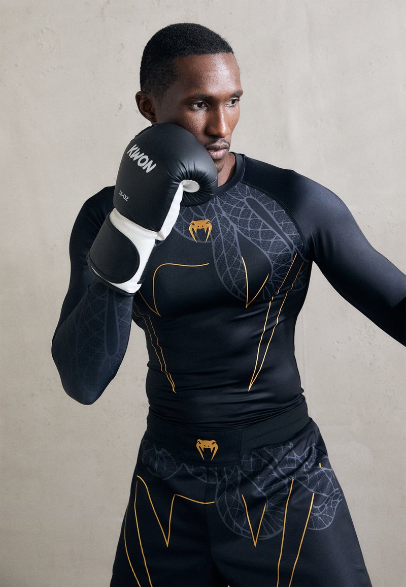 T-shirt de sport noir à manches longues avec un motif de serpent et des accents jaunes. Shorts noirs avec un design similaire. Modèle portant des gants de boxe noirs.