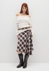 Suéter blanco con hombros al aire, falda a cuadros en tonos marrones, cinturón marrón con detalles plateados y botas negras hasta la rodilla con correas.
