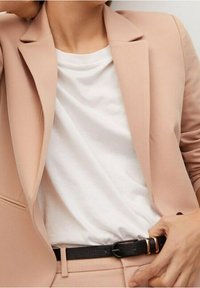 Béžový blazer s hladkou texturou, zkombinovaný s bílým tričkem. Viditelný je také černý opasek se zlatou sponou.