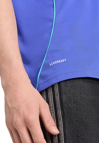 Nahbillede af en hånd, der hviler nær hoften, iført en blå skjorte med teksten "AEROREADY" og mørke bukser med tre lodrette striber.