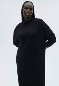 Abito nero in maglia con scollatura alta, maniche ampie e una vestibilità rilassata. Il tessuto appare morbido e lavorato, adatto per un abbigliamento casual.