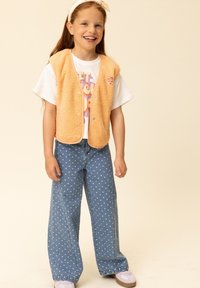 Oranje fleecevest met knoopsluiting over een wit T-shirt met een grafische print. Blauwe jeans met wijde pijpen en witte hartjespatronen. Lichte sneakers.