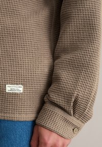 Bruine textuurrijke lange mouwen shirt met een ruitpatroon, voorzien van een geruite manchetknoop en een klein merkembleem op de mouw. Denim eronder.