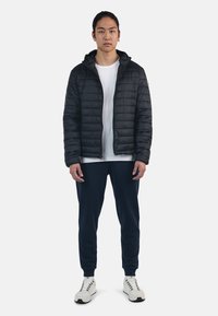 Giacca nera imbottita con zip e cappuccio, indossata sopra una maglietta bianca. Abbinata a pantaloni jogging blu navy e sneaker bianche. Tessuto liscio, design casual.