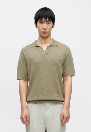 Πράσινο ελιάς κοντομάνικο πόλο με υφή, V-λαιμόκοψη και μικρό γιακά. Το ύφασμα φαίνεται πλεκτό ή βρόχινο, δημιουργώντας βάθος και διάσταση.