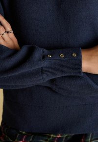 Marineblauer Strickpullover mit gerippten Bündchen und drei Akzenten aus goldenen Knöpfen. Die Textur ist weich und der Schnitt wirkt lässig.