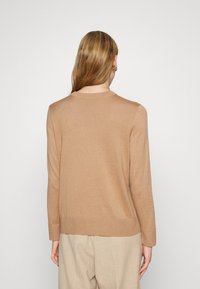Femme aux cheveux longs châtain clair, portant des boucles d'oreilles créoles dorées, un pull couleur camel et un pantalon beige, debout face à un fond blanc.