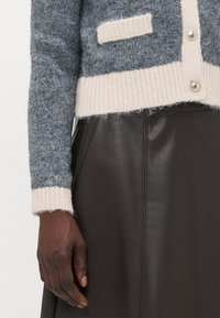 Cardigan grigio a maglia con orlo e polsini a costine beige chiaro, dotato di chiusura con bottoni e di una piccola tasca sul petto, abbinato a una gonna in pelle nera.