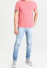 Man som bär en rosa t-shirt med kort ärm, ljusblå jeans, grå högskaftade sneakers, med en tatuering på vänster arm, stående mot en vit bakgrund.