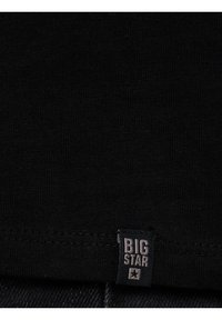 Gros plan sur un tissu noir avec une petite étiquette rectangulaire portant l'inscription « BIG STAR » et une icône d'étoile cousue sur l'ourlet inférieur.