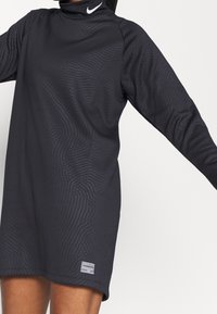 Mustat pitkähihaiset mekko, jossa on korkea kaulus, hienovarainen aaltoilu kuvio ja pieni Nike-logo vasemmassa olkapäässä. Puuvillasekoitekangas.