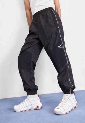 Osoba w czarnych spodniach dresowych Nike Air i białych butach Nike Air z czerwonymi akcentami, stojąca na niebieskiej podłodze na tle białej ściany.