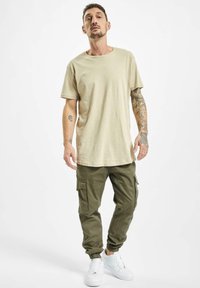 Bežové tričko a olivové cargo nohavice s vreckami. Biele tenisky. Voľný strih s okrúhlym výstrihom a elastickými manžetami na nohaviciach.