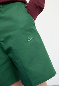 Calções de algodão verdes com bolsos, apresentando um logótipo da Nike bordado em verde mais escuro. O tecido tem uma textura suave e um design estruturado.