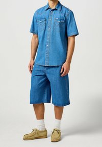 Ljusblå denimskjorta med kort ärm, tryckknappar och två bröstfickor, ihop med matchande denimshorts och beige slip-on-skor.