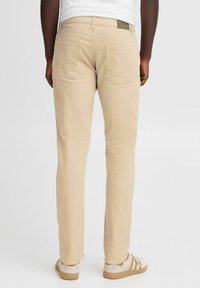 Pantaloni in cotone beige con vestibilità slim, dotati di due tasche sul retro e un'etichetta in pelle sulla vita. Indossati con scarpe beige e bianche.