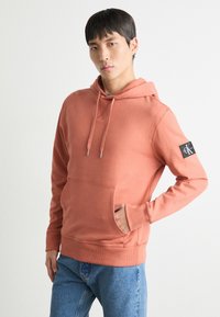 Felpa con cappuccio arancione, dotata di tasca frontale e polsini a coste. Presenta un'etichetta con il marchio sulla manica sinistra. Indossata sopra un paio di jeans blu.