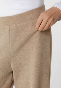 Beige Strickhosen mit einer weichen Textur und elastischem Bund; eine Hand greift nach der Seitentasche, die den praktischen Nutzen und das Design betont.