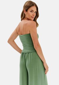 Zielony, bezramnowy kombinezon wykonany z miękkiego, elastycznego materiału o luźnym kroju i zebranym pasie. Posiada gładką teksturę i łatwo się układa.