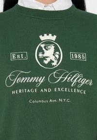 Felpa verde con collo a costine. Presenta ricami bianchi: stemma del leone, "Tommy Hilfiger", "Est. 1985", e "ERITAGE AND EXCELLENCE."