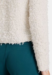 Pull en peluche blanc avec une coupe cropped, mettant en avant une finition brute et des manches longues, associé à un pantalon taillé teal.