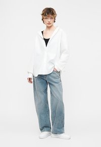 Γυναίκα που φοράει υπερμεγέθη λευκό πουκάμισο με κουμπιά πάνω από μαύρο τοπ, χαλαρά ανοιχτόγαλάζια τζιν και λευκά αθλητικά, στέκεται με το ένα χέρι στην τσέπη.