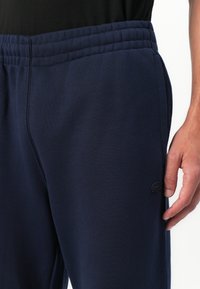 Mann som har på seg marineblå joggebukser med elastisk midje og en liten svart brodert logo nær lommen.