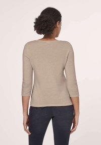 Frau mit lockigem, zurückgebundenem Haar, die ein beiges langärmliges Oberteil und dunkle Jeans trägt und von einer neutralen Hintergrundfarbe abgewandt steht.