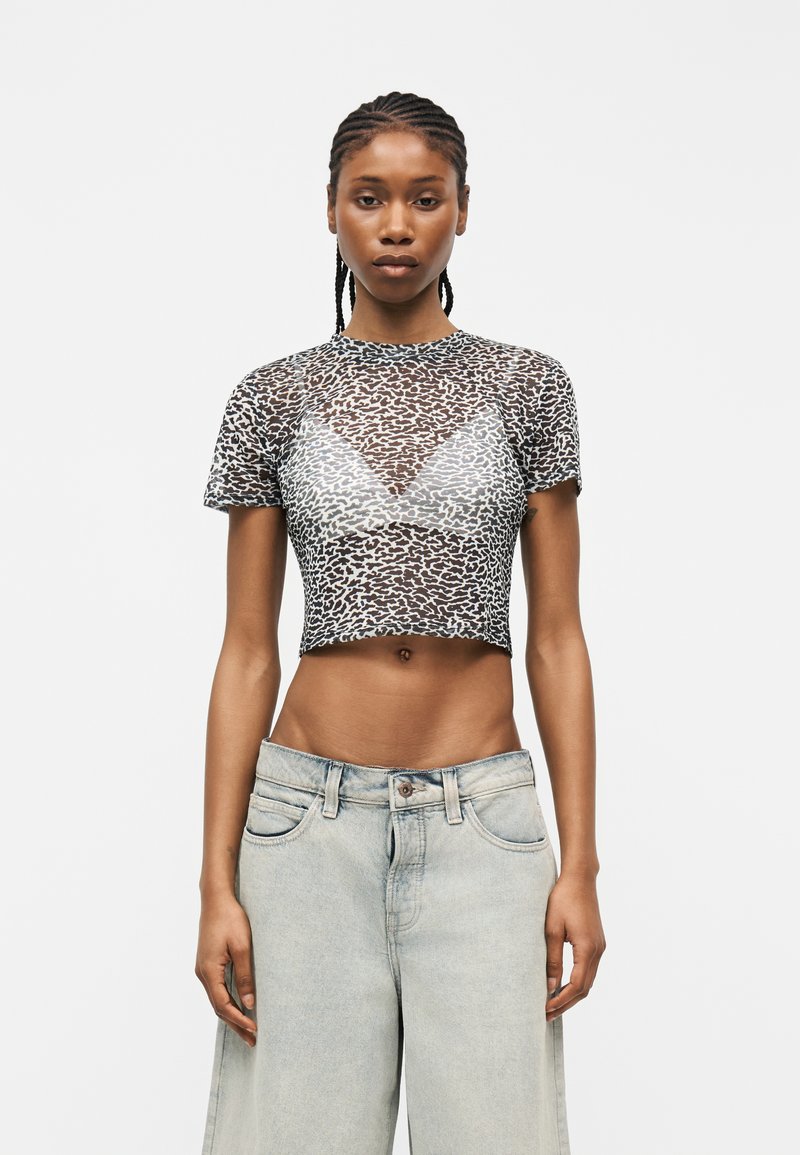 Γυναίκα με πλεξούδες, που φοράει see-through crop top με λεοπάρ σχέδιο πάνω από λευκό σουτιέν και χαλαρό ανοιχτό μπλε τζιν, που στέκεται μπροστά σε απλό φόντο.