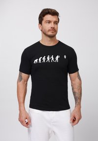 Black short-sleeved t-shirt featuring a white graphic depicting evolution. Crew neckline and fitted design. Subtle logo on the right.